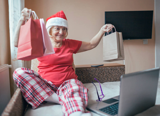 Festeggiamenti di Natale online sicuri. Donna anziana in cappello rosso Santa dando regalo alla sua famiglia praticamente con internet e notebook. Videochiamata. Soggiorno a casa, vacanza lontana, stile di vita interno emotivo - Foto, immagini