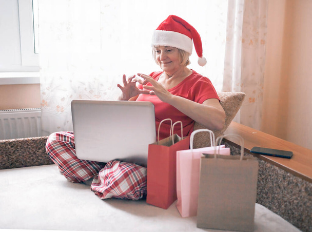 Turvallinen online joulujuhla. Vanhempi nainen Santa punainen hattu antaa lahjaksi perheelleen lähes internetin ja muistikirjan. Videopuhelu. Pysy kotona, kaukainen loma, emotionaalinen sisäelämäntapa - Valokuva, kuva