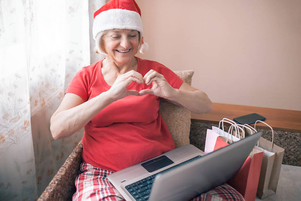 Safe online Christmas celebration. Senior woman in Santa red hat giving present to her family virtually with internet and notebook. Video call. Stay home, distant holiday, emotional indoor lifestyle - Photo, Image