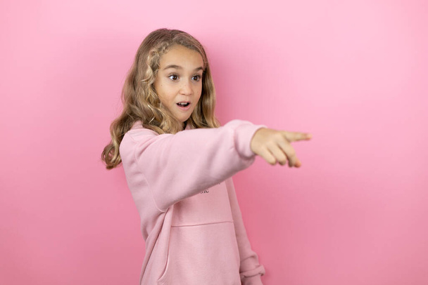 Молода красива дівчинка стоїть над ізольованим рожевим тлом, вказуючи пальцем здивований попереду, відкритий рот здивований вираз, щось на передньому плані
 - Фото, зображення