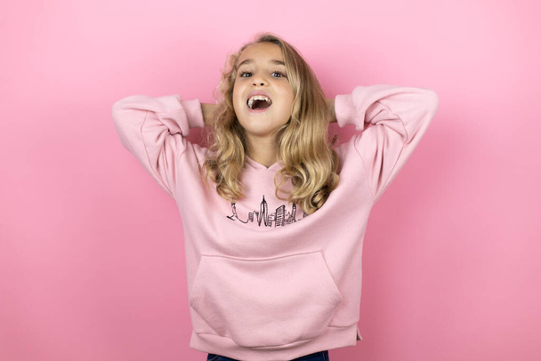 Fiatal gyönyörű gyermek lány áll az elszigetelt rózsaszín háttér pihentető és nyújtás, karok és kezek a fej és a nyak mögött mosolyogva boldog - Fotó, kép
