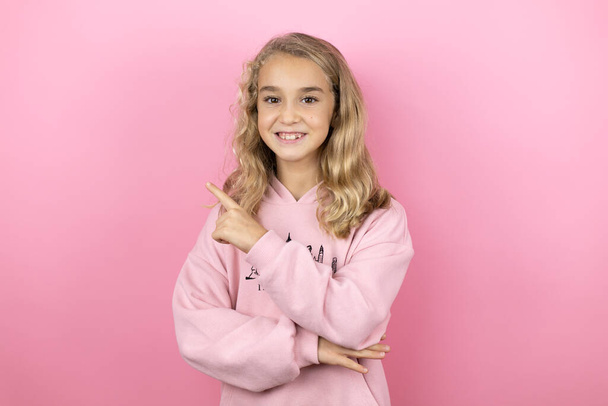 Young beautiful child girl standing over isolated pink background smiling confident pointing with hand and finger to the side - Photo, Image