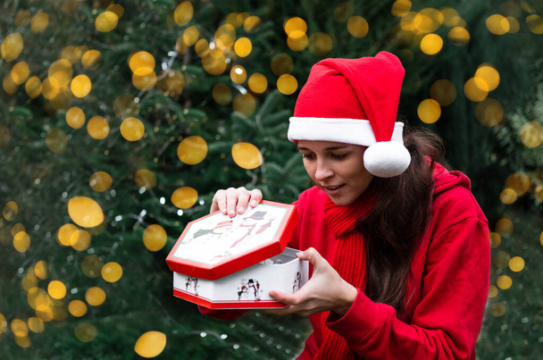 Söpö eurooppalainen nuori nainen punaisessa Santa hatussa avaa joululahjan yllättynyt ja onnellinen. Kirkkaan keltaisia sumeita valoja seppeleestä. Vastaanottaa uuden vuoden lahja. - Valokuva, kuva