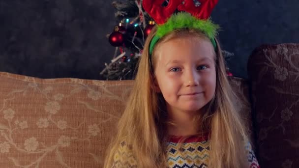 Happy little girl in red deer horns laughs and looks into camera at home 4K face portrait. Smiling toothless child in New Year eve Christmas tree lights. Family holidays celebration spirit fun concept - Footage, Video