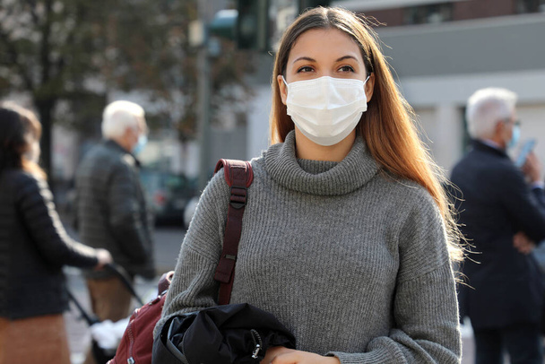 Portrait of young student woman walking in city street wearing surgical mask. Girl with face mask walks on road and people respecting social distancing stopped at the traffic lights on background. - Photo, image