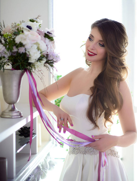 A beautiful smiling bride with wedding makeup. Wedding morning in a white dress and with a bouquet in the interior. - Foto, imagen
