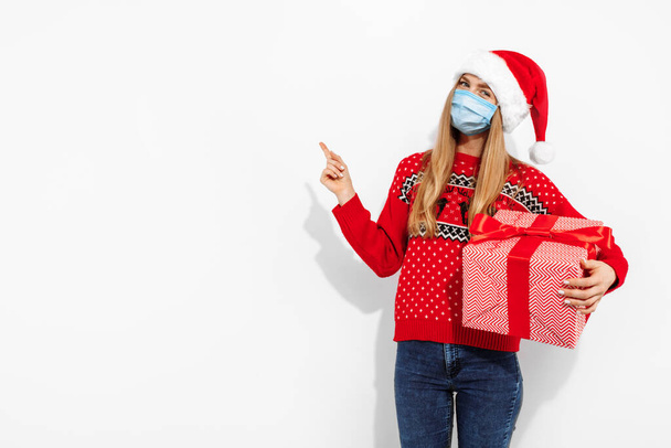 Smiling attractive young woman wearing christmas santa claus hat and medical protective mask on face to protect from viruses, holding gift box and pointing to copyspace on white background - Photo, image