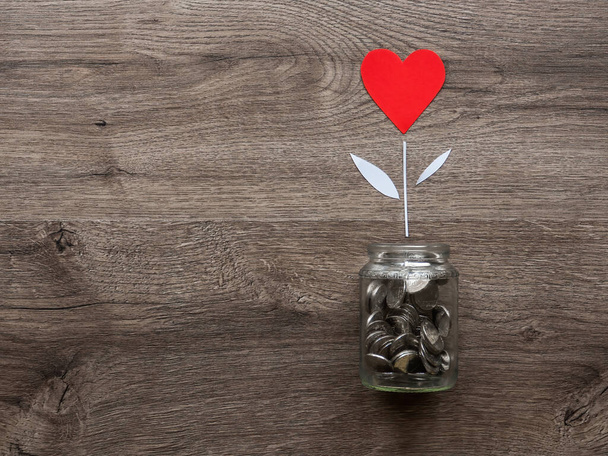 Una lata de vidrio con monedas de metal y una flor con corazón rojo creciendo en ella. Fondo de madera. Vista superior - Foto, imagen