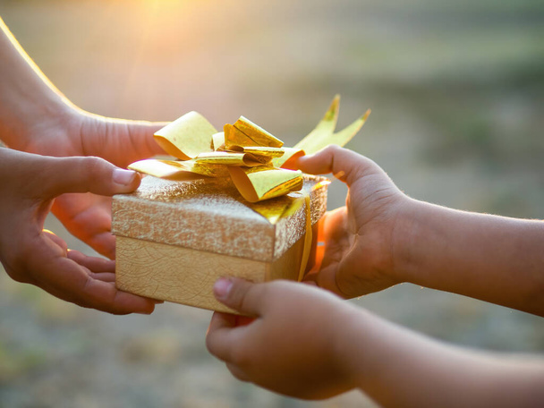 Un niño y un niño sostienen una caja de regalo con cinta dorada, poniéndose el sol. Concepto de vacaciones - Foto, Imagen