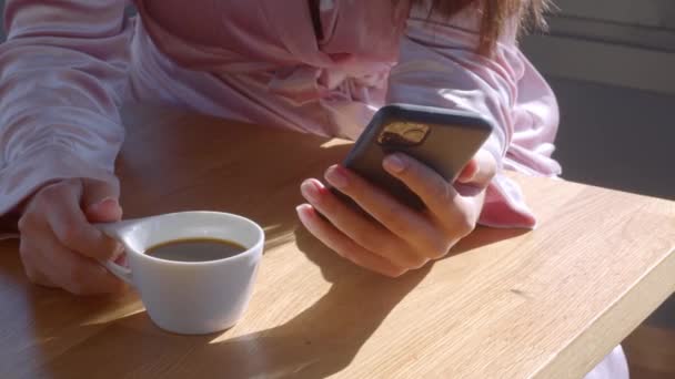Közelről, arc nélkül. Fehér lány, rózsaszínű köntösben, kezében egy telefonnal, iszik egy csésze kávénál a napsütéses reggelen. Panned video kiváló minőségű 4k felvételek. - Felvétel, videó