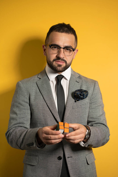 Smart young bearded handsome man in eye glasses wearing a suite holds a solved Pocket Cube 2x2x2 rotatable puzzle in hands while showing it on camera. Isolated on yellow background - Fotó, kép