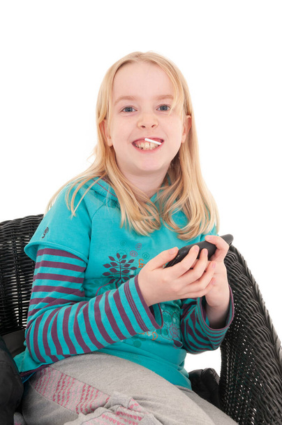 Gros plan d'une jeune fille blonde heureuse assise sur une chaise en osier tenant un smartphone. Isolé sur fond de studio blanc - Photo, image