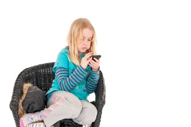 Jong blond meisje zat op een rieten stoel met een smartphone. Geïsoleerd op witte studio achtergrond - Foto, afbeelding