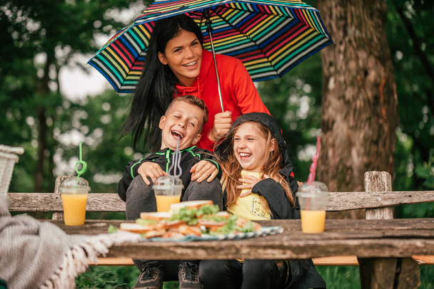 Beautiful mother with two kids are sitting at a picnic table in the forest. Happy family having fun together, mother holding umbrella while it is raining. Selective focus - Photo, Image