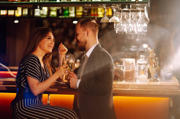 Young happy couple in love toasting with champagne on the bar. Smiling man and woman clinking a glass champagne, enjoying winter holiday. Party, celebration, drink, birthday concept. - Zdjęcie, obraz