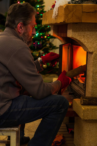 New Year by the fireplace. A man with a beard lays a log in the burning fireplace. In the background, New Year lights are lit on the tree - Foto, Imagen