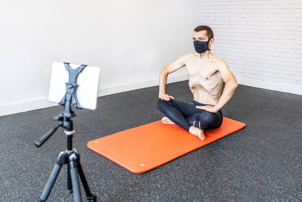 caucasian man with a face mask training hypopressive abdominal exercises on the mat, connected to his tablet performing online class for his students - Фото, изображение
