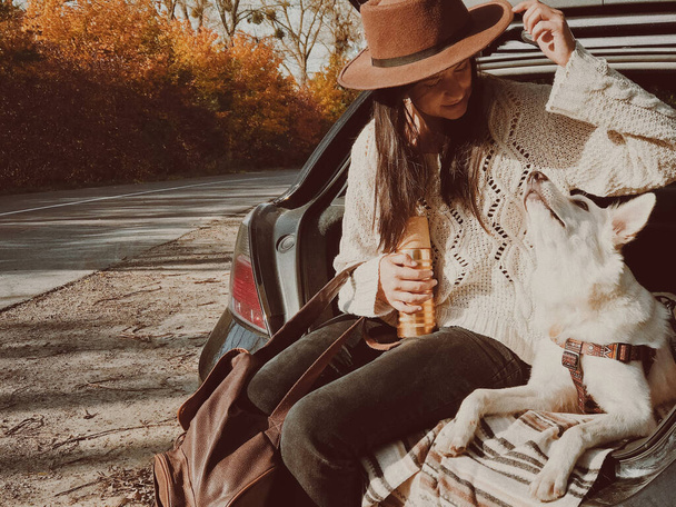 Stylová mladá žena sedí se svým bílým psem v kufru auta na pozadí podzimních stromů. Výlet s domácím mazlíčkem v podzimní sezóně. Žena cestuje sama se štěňátkem a prozkoumává zemi - Fotografie, Obrázek