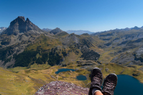 Botas enfocadas de un excursionista en la cima del pico Ayous con vistas desenfocadas del lago de montaña Ayous y las montañas de los Pirineos con el Midi d 'Ossau en frente. - Foto, Imagen