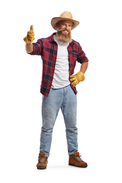 Full length portrait of a cheerful bearded farmer with gloves and a straw hat showing thumbs up isolated on white background - Foto, Imagen