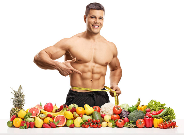 Topless muscular man measuring waist and posing with healthy fruits and vegetables isolated on white background - Foto, Bild