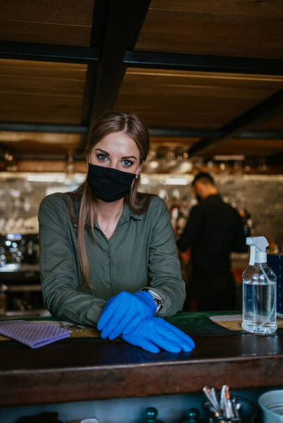 Ritratto di una cameriera seria in piedi dietro un bar in un bel ristorante. Indossa una maschera protettiva e guanti come parte delle misure di sicurezza contro la pandemia di Coronavirus. - Foto, immagini