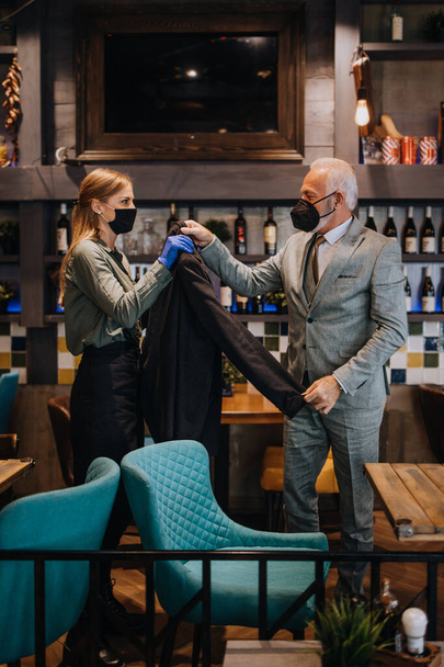 Confident senior businessman standing in exclusive restaurant. He takes off his coat while the waitress helps him. They both wearing protective face masks against virus infection. - Zdjęcie, obraz