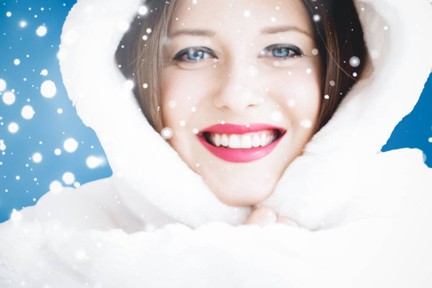 Happy Christmas and winter holiday portrait of young woman in white hooded fur coat, snow on blue background, fashion and lifestyle - Photo, Image