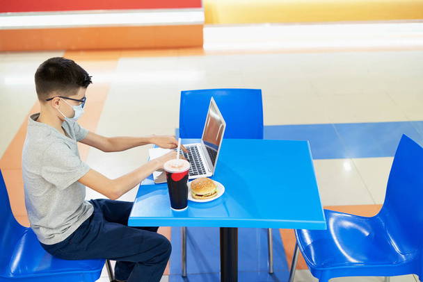 Teenager in fast food restaurant wearing protective mask with laptop - Φωτογραφία, εικόνα