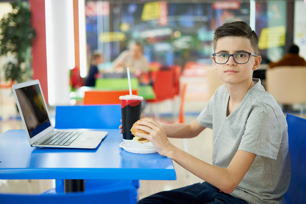 Portrait d'adolescent attrayant avec hamburger et ordinateur portable à la cour alimentaire du centre commercial - Photo, image