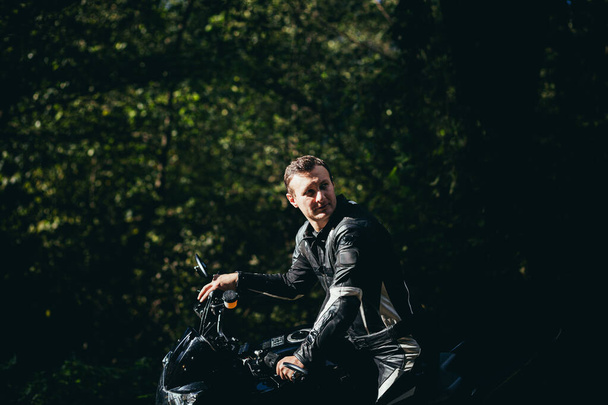 A man on a black sports motorcycle in black leather clothes on the road in the woods - Photo, Image