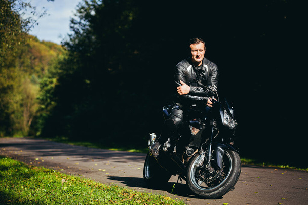 A man on a black sports motorcycle in black leather clothes on the road in the woods - Foto, Bild