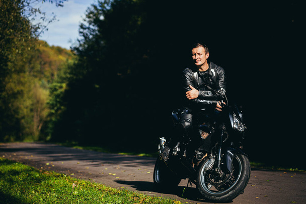 A man on a black sports motorcycle in black leather clothes on the road in the woods - Foto, imagen