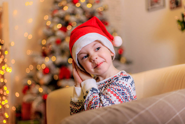 パジャマ姿の男の子と木の下のサンタの帽子。男の子とクリスマスツリー。可愛い子ね。休日だ。新年とクリスマスについての記事。カラフルなトウヒ。ガーランド。休日の装飾と装飾 - 写真・画像