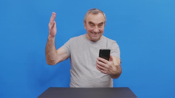 Happy grandfather winner mobile app sport bet holding smartphone having positive emotion. Overjoyed elderly man laughing celebrating victory success read good news message. Isolated on blue. - Filmati, video