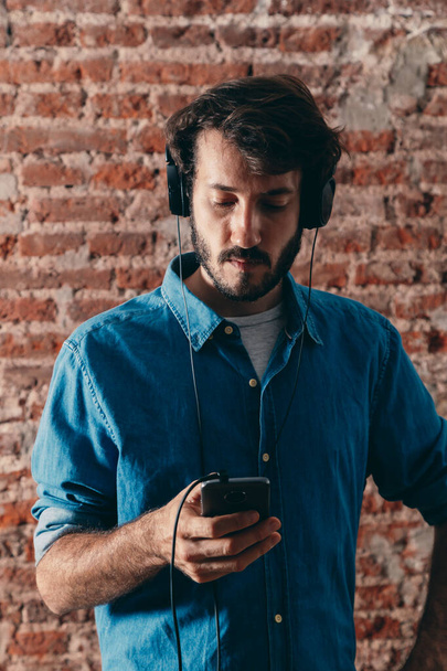 Retrato de un joven con auriculares y teléfono celular. Ropa casual y pared de ladrillo de fondo. - Foto, imagen