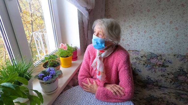 Zamknięte. Covid-19, koncepcja zdrowia, bezpieczeństwa i pandemii - starsza samotna kobieta nosząca ochronną maskę medyczną siedząca w pobliżu okna w domu dla ochrony przed wirusem - Zdjęcie, obraz
