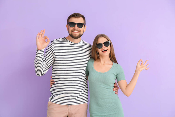 Νεαρό ζευγάρι με κομψά γυαλιά ηλίου που δείχνει OK χειρονομία στο φόντο χρώμα - Φωτογραφία, εικόνα