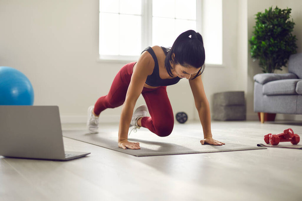 Sporty young woman watching video lesson and doing plank with knee drive at home - Fotoğraf, Görsel
