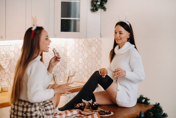 two girls in a cozy home environment in the kitchen with champagne in their hands for Christmas. Smiling girls drink champagne on a festive evening. - Foto, afbeelding
