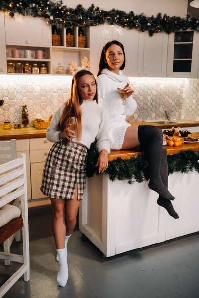 two girls in a cozy home environment in the kitchen with champagne in their hands for Christmas. Smiling girls drink champagne on a festive evening. - 写真・画像