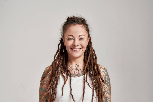 Portrait of young tattooed caucasian woman with dreadlocks wearing white shirt, smiling at camera while posing isolated over grey background - Foto, immagini