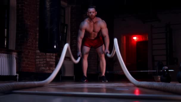 Hombre duro sin camisa de pie en el gimnasio y haciendo ejercicios de cuerdas de batalla - Metraje, vídeo