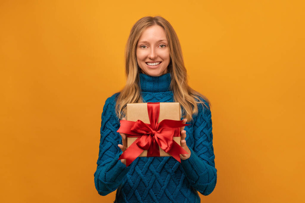 Зображення чарівної молодої жінки в теплому в'язаному светрі посміхається і тримає подарунок з червоною стрічкою. Студійні зйомки, жовтий фон. Новий рік, жіночий день, день народження, святкова концепція - Фото, зображення
