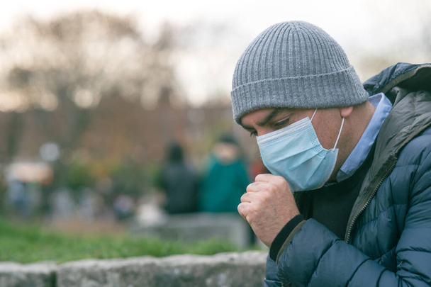 Πρόληψη της εξάπλωσης της νόσου του Coronavirus 2019 (COVID-19). Ο άνθρωπος στο πάρκο φτερνίζεται ή βήχει σε μια προστατευτική ιατρική μάσκα προσώπου - Φωτογραφία, εικόνα