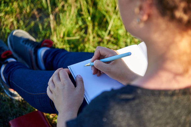 Frau schreibt mit Stift in sauberes Notizbuch im Gras in der Natur. - Foto, Bild