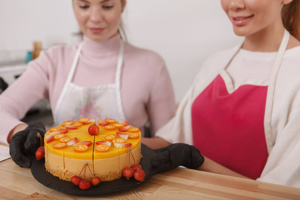 Cropped shot of two female confectioners garnishing and decorating delicious raw mango vegan cake, working at their bakery - Fotografie, Obrázek