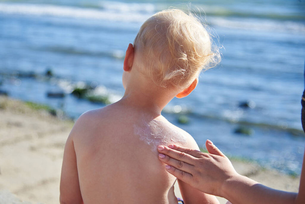 Moeder smeert haar zoontje terug met zonnebrandcrème op het strand. - Foto, afbeelding