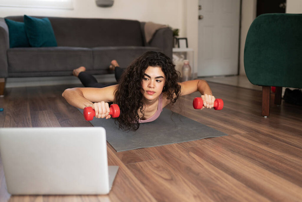 Beautiful latin young woman watching an online workout tutorial on her laptop while exercising with dumbbells in a locust pose at home - Foto, immagini