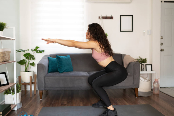 Vista lateral de una joven latina entrenando y haciendo ejercicio en casa. Mujeres atractivas de 20 años haciendo sentadillas en su sala de estar - Foto, imagen